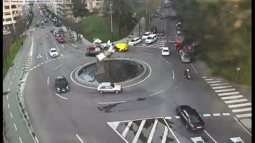 A roundabout with several cars and a scooter, surrounded by roads and pedestrian walkways on an overcast day.