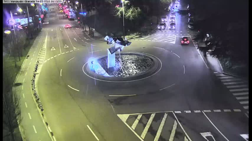 A nighttime aerial view shows a roundabout with a modern sculpture in the center, surrounded by roadways with sparse traffic.