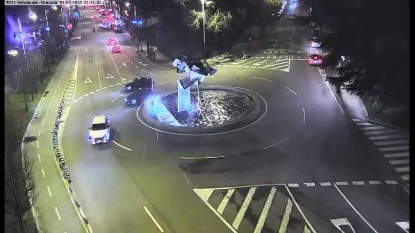 Several cars are driving around a roundabout at night under artificial light.