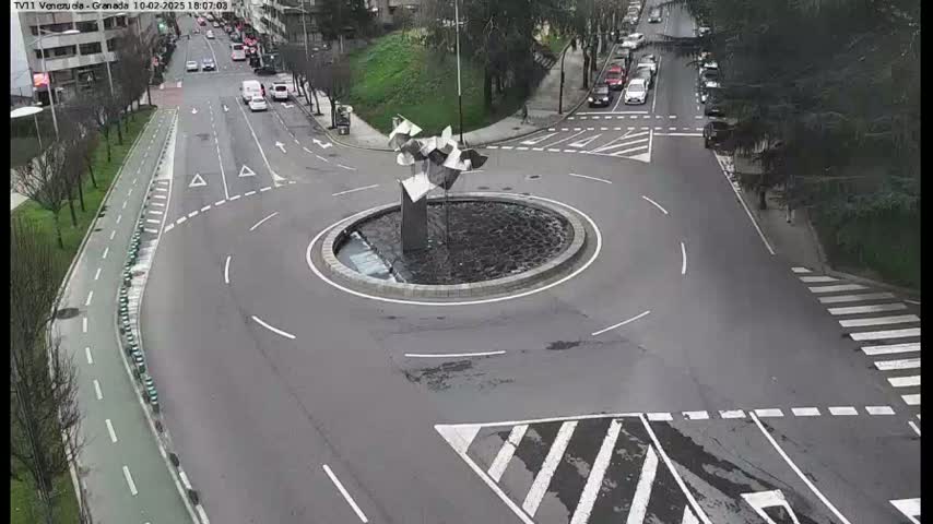 A traffic circle with a modern sculpture in the center is surrounded by roads and a bike lane, all under a seemingly overcast sky.