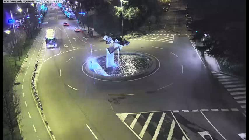 A nighttime aerial view shows a roundabout with a modern sculpture at its center, surrounded by streets with sparse vehicle traffic.
