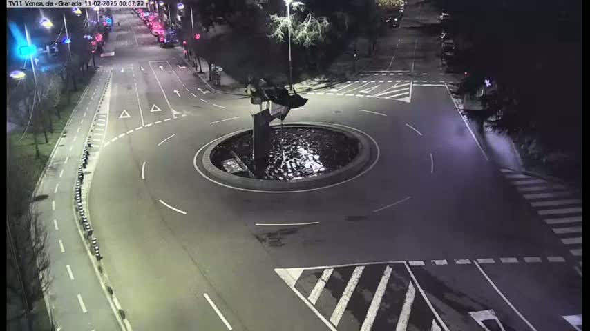 A nighttime aerial view shows a traffic circle with a fountain in the center, surrounded by roadways and pedestrian crosswalks.