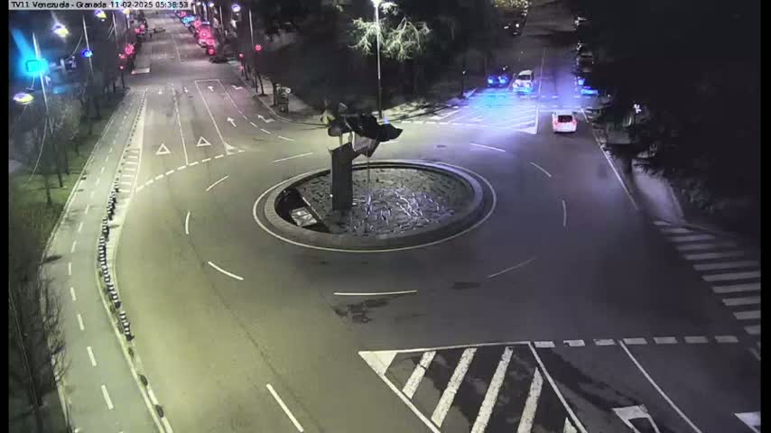 A nighttime overhead view of a roundabout with a few cars driving around it and adjacent roads.