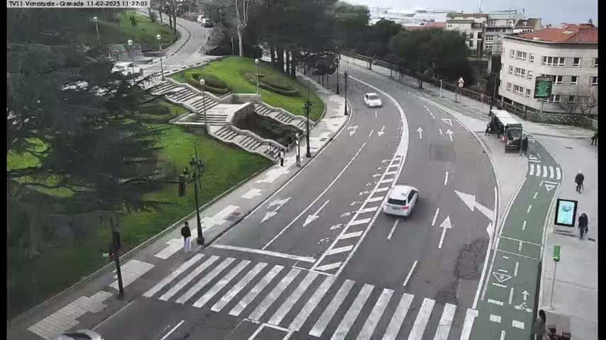 A curved road with cars and pedestrians, next to a grassy area with stairs and trees, on an overcast day.