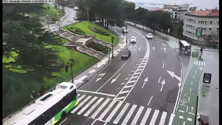 A wet road curves through a park-like setting, with a bus and several cars visible, alongside a pedestrian crossing and bike lane.