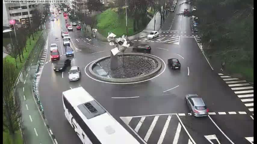 A wet roundabout with several cars, a bus, and a modern sculpture in the center.