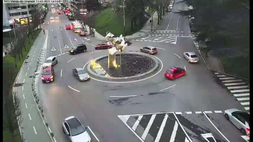 Several cars navigate a roundabout in the center of a road intersection, under overcast daylight conditions.