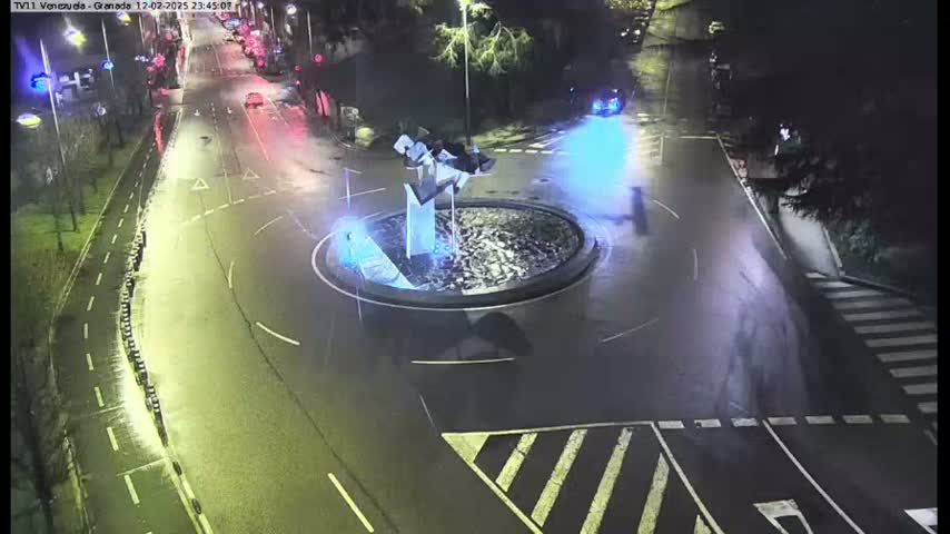 A wet roundabout with a modern sculpture in the center is surrounded by nighttime traffic on a rain-slicked road.