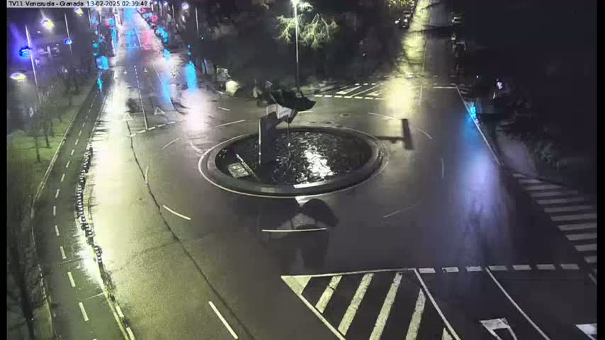 A nighttime aerial view shows a wet roundabout with a fountain at its center, surrounded by streets and crosswalks.