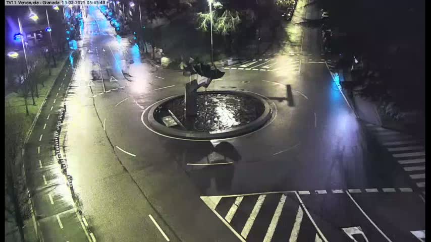 A nighttime aerial view shows a circular traffic island with a fountain in the center of a wet road intersection.
