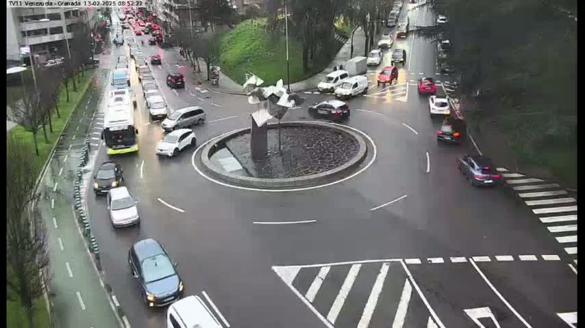 A wet roundabout in a city is surrounded by cars, with several parked on the side of the road and others navigating the circle.