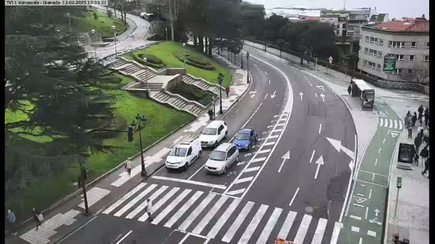A curved road with several white cars and a pedestrian crossing, situated beside a grassy area with stairs and trees, under overcast skies.