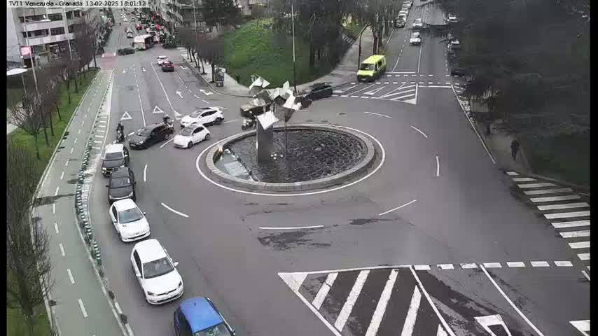 A roundabout with several cars, a modern sculpture in the center, and a pedestrian crossing, on an overcast day.