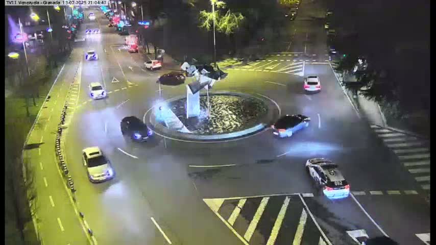 Several cars are driving around a circular traffic island with a fountain at its center at night.