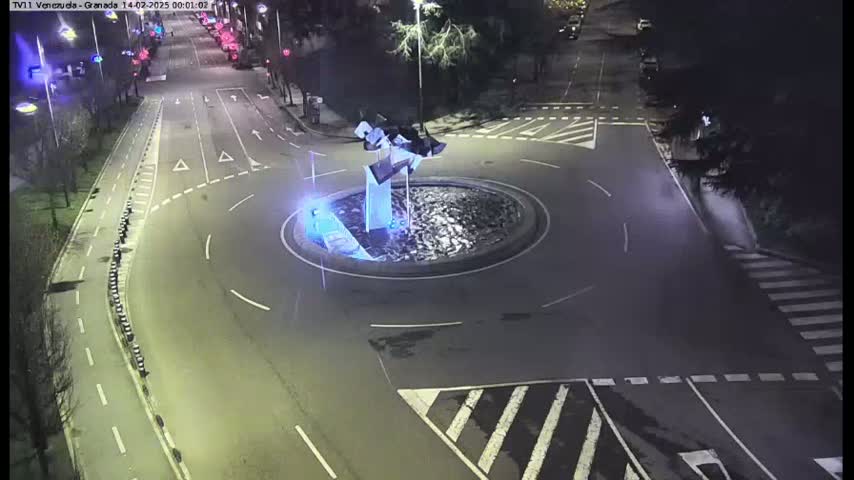 A nighttime aerial view shows a traffic circle with a modern sculpture in the center, surrounded by roads with sparse traffic and pedestrian crossings.