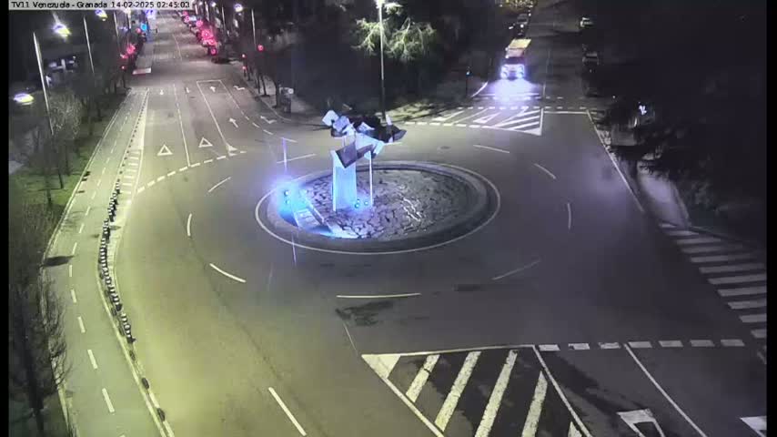 A nighttime aerial view shows a roundabout with a modern sculpture in the center, surrounded by roads and pedestrian crossings.