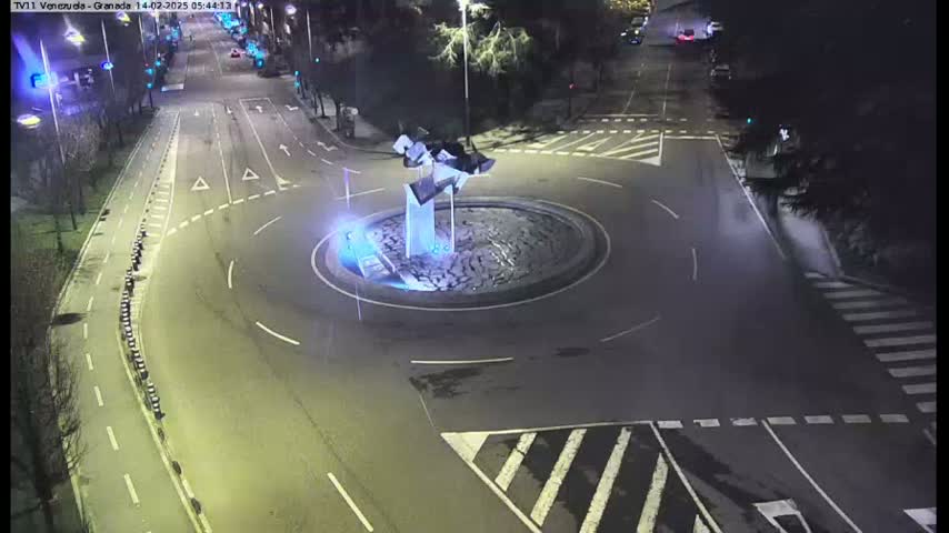 A nighttime aerial view of a roundabout with a modern sculpture in its center, surrounded by roads and pedestrian crossings.