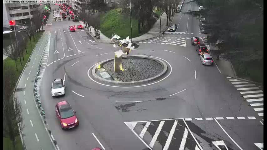 An overcast day shows several cars navigating a roundabout with a modern metal sculpture in the center.