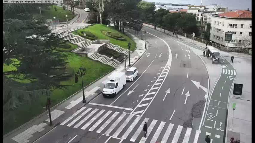 A curving road with a pedestrian crossing and bike lane is seen from above, with a few vehicles and pedestrians present on an overcast day.