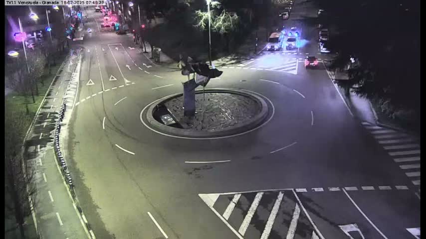 A nighttime aerial view of a roundabout with a sculpture in the center, surrounded by roadways and some parked vehicles.