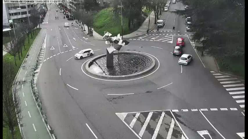 A roundabout with several cars and a modern sculpture in the center, surrounded by streets with pedestrian crossings and bike lanes, on an overcast day.