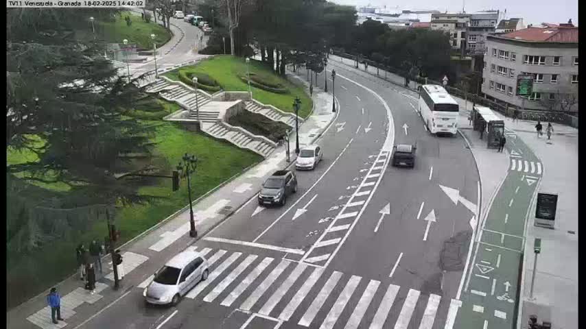 A curved road with several cars and buses passes through a city park with green lawns, stairs, and trees on an overcast day.