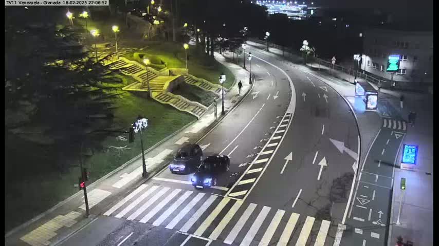 Two dark-colored cars are stopped at a crosswalk on a well-lit roadway at night.