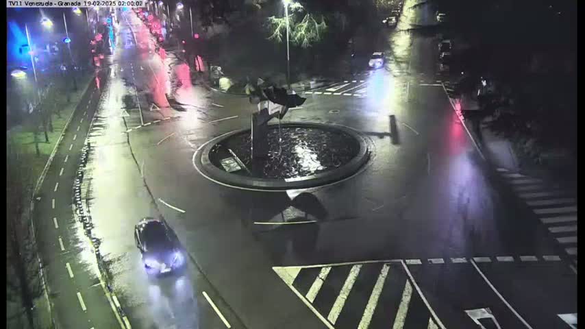 A wet, nighttime scene shows a roundabout with a fountain at its center, surrounded by roadways and crosswalks with a few cars driving around the area.