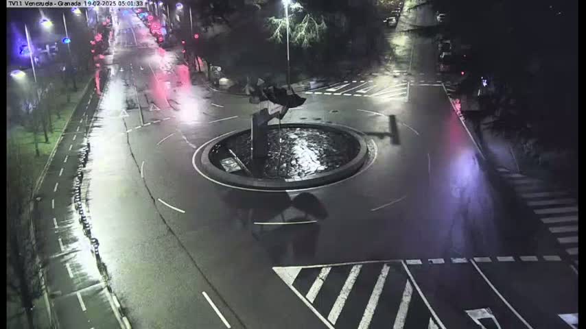 A nighttime aerial view of a wet roundabout with sparse traffic and a sculpture in the center.