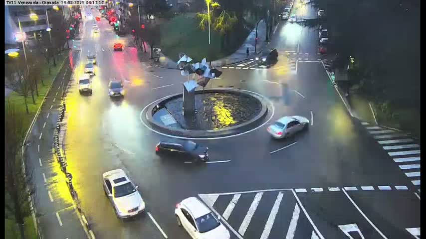 A wet roundabout with several cars navigating it, surrounded by roads with additional vehicles, under a dark, overcast sky.
