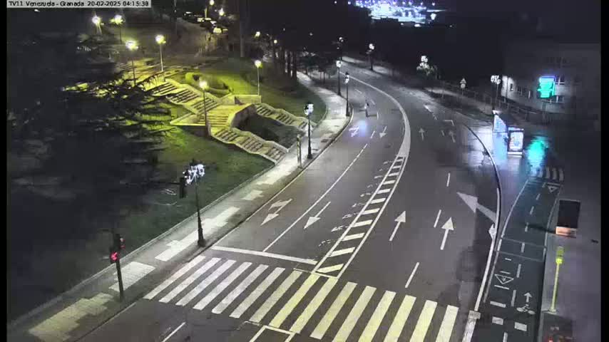 A nighttime, high-angle view shows a mostly empty road curving through a park-like area with stairs and trees, under light rain.