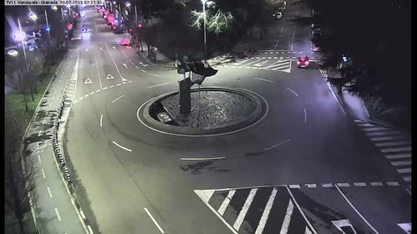 A nighttime aerial view shows a roundabout with a dark sculpture in the center, surrounded by a roadway with several cars and pedestrian crossings.