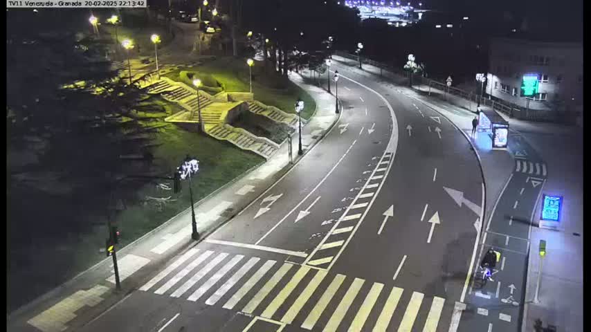 A nighttime, brightly lit, curved roadway with crosswalks and a bicycle lane next to a grassy area with stairs, under clear skies.