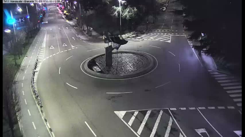 A nighttime, overhead view shows an empty roundabout with a dark, sculptural centerpiece in the middle of a road intersection.