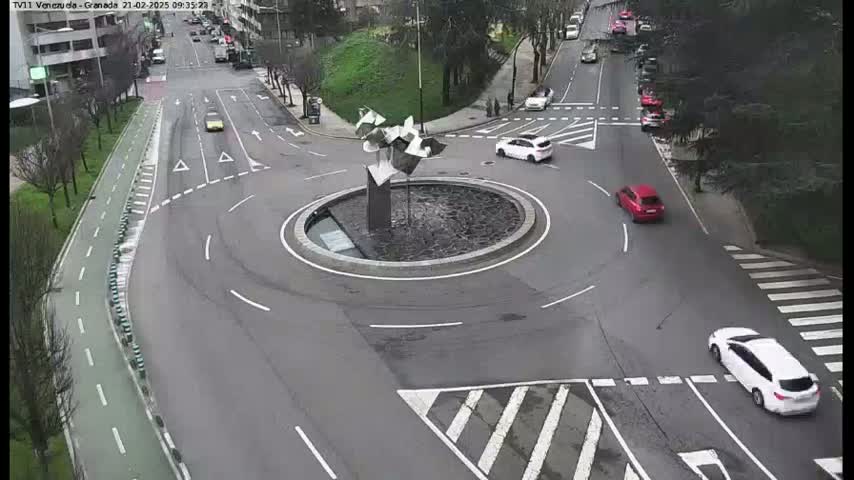 An aerial view shows a roundabout with several cars, a modern sculpture in the center, and a pedestrian crossing.