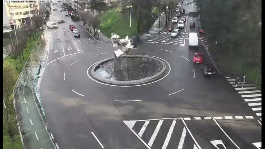 An aerial view of a roundabout with several cars and a pedestrian crossing, on a wet day.