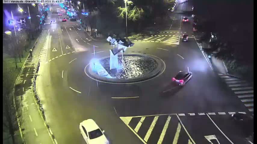 A nighttime aerial view shows a roundabout with a modern sculpture in the center, several cars navigating the roadway, and wet pavement.