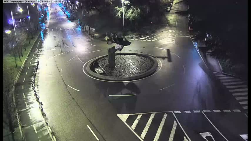 A nighttime overhead view shows a roundabout with a dark sculpture in the center, surrounded by dark streets and crosswalks.
