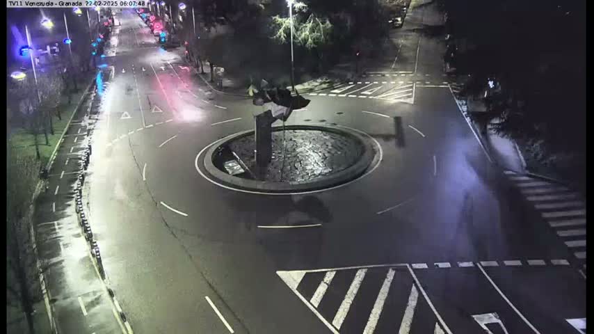 A wet, nighttime aerial view shows a roundabout with a sculpture in the center, surrounded by roads and pedestrian crossings.