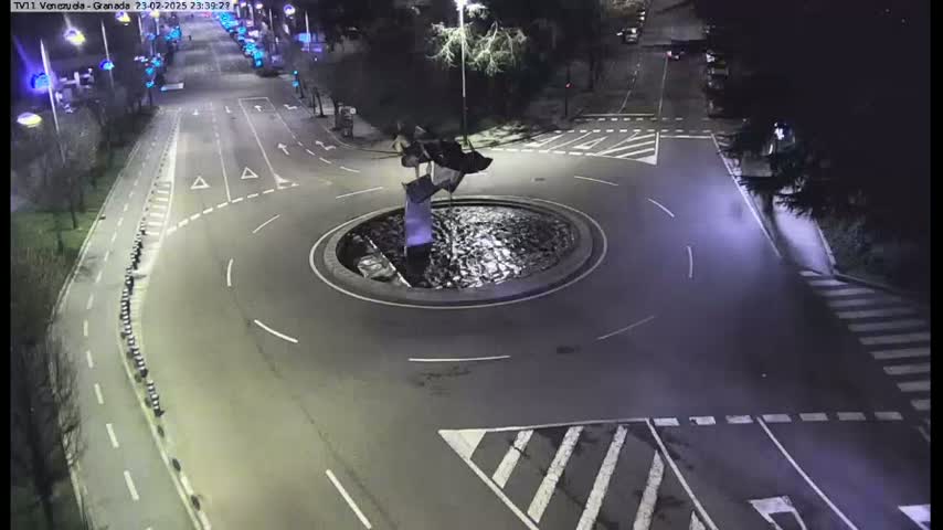 A nighttime aerial view of a roundabout with a modern sculpture in the center, surrounded by roads and pedestrian crossings.