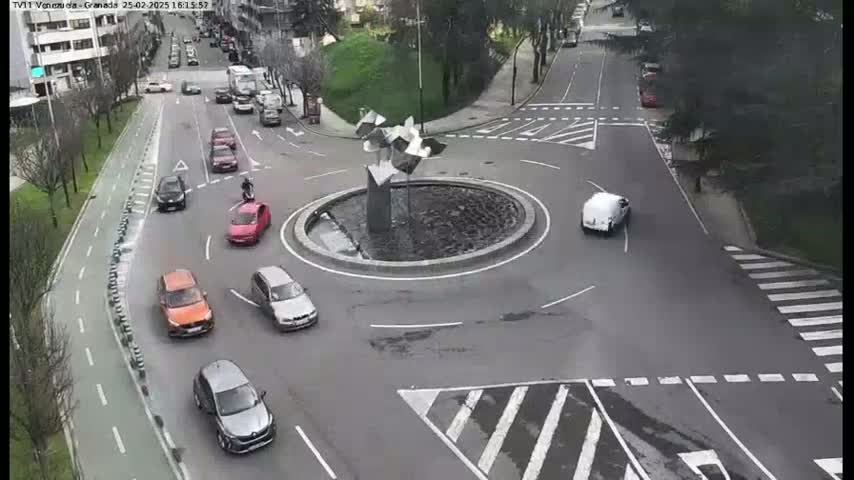 Several cars are driving around a roundabout with a modern metal sculpture in the center, on a partly cloudy day.