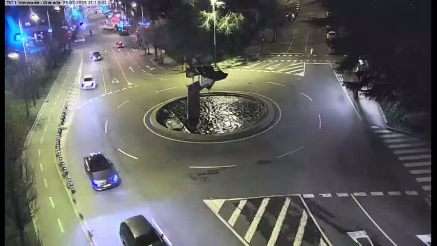 A nighttime aerial view shows a roundabout with several cars driving around it and along intersecting roads, with streetlights illuminating the scene.