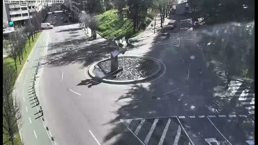 A traffic circle with a fountain in the center is surrounded by roads, a bike lane, and trees, under a sunny sky.