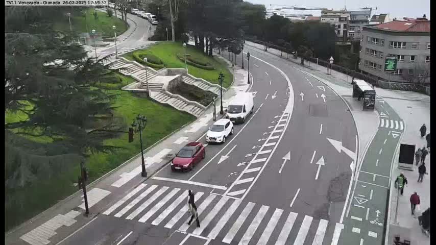 A pedestrian crosses a crosswalk on a curving road with several cars and a van parked alongside,  under an overcast sky.