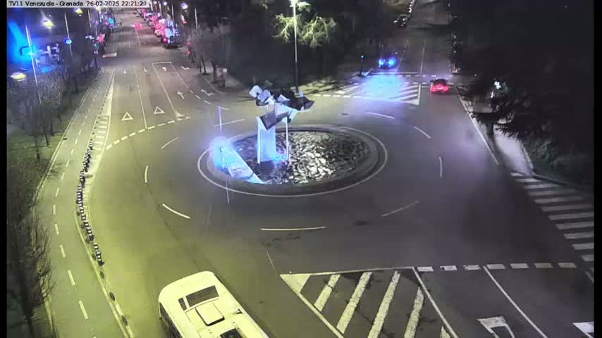 A nighttime aerial view shows a roundabout with a modern sculpture at its center, surrounded by roads and several vehicles, including a bus.