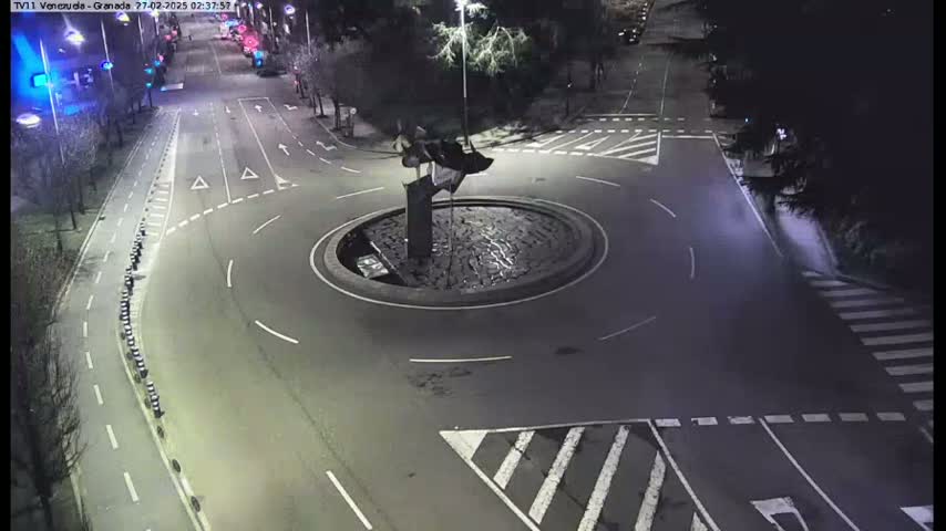 A nighttime aerial view shows a roundabout with a dark sculpture in the center, surrounded by roadways and pedestrian crosswalks.