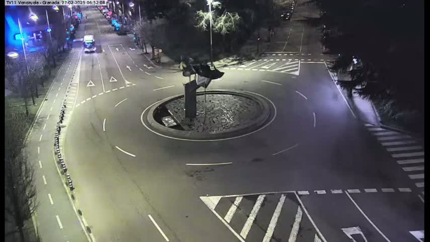 A nighttime aerial view shows a roundabout with a dark sculpture in the center, surrounded by roadways with sparse traffic.