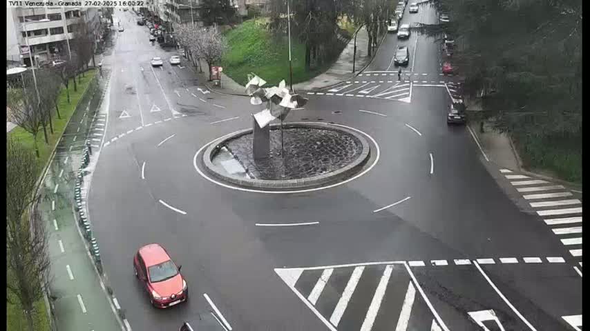 A wet roundabout with a modern sculpture in the center, surrounded by roads with several cars and a pedestrian crossing.
