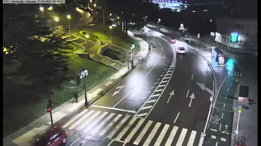 A wet, nighttime street scene shows a curving road with a pedestrian crossing, several cars, and streetlights illuminating the area.