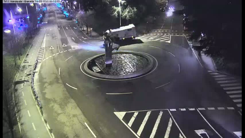 A nighttime aerial view shows a roundabout with a central fountain, a white van, and several street markings and crosswalks on surrounding roads.